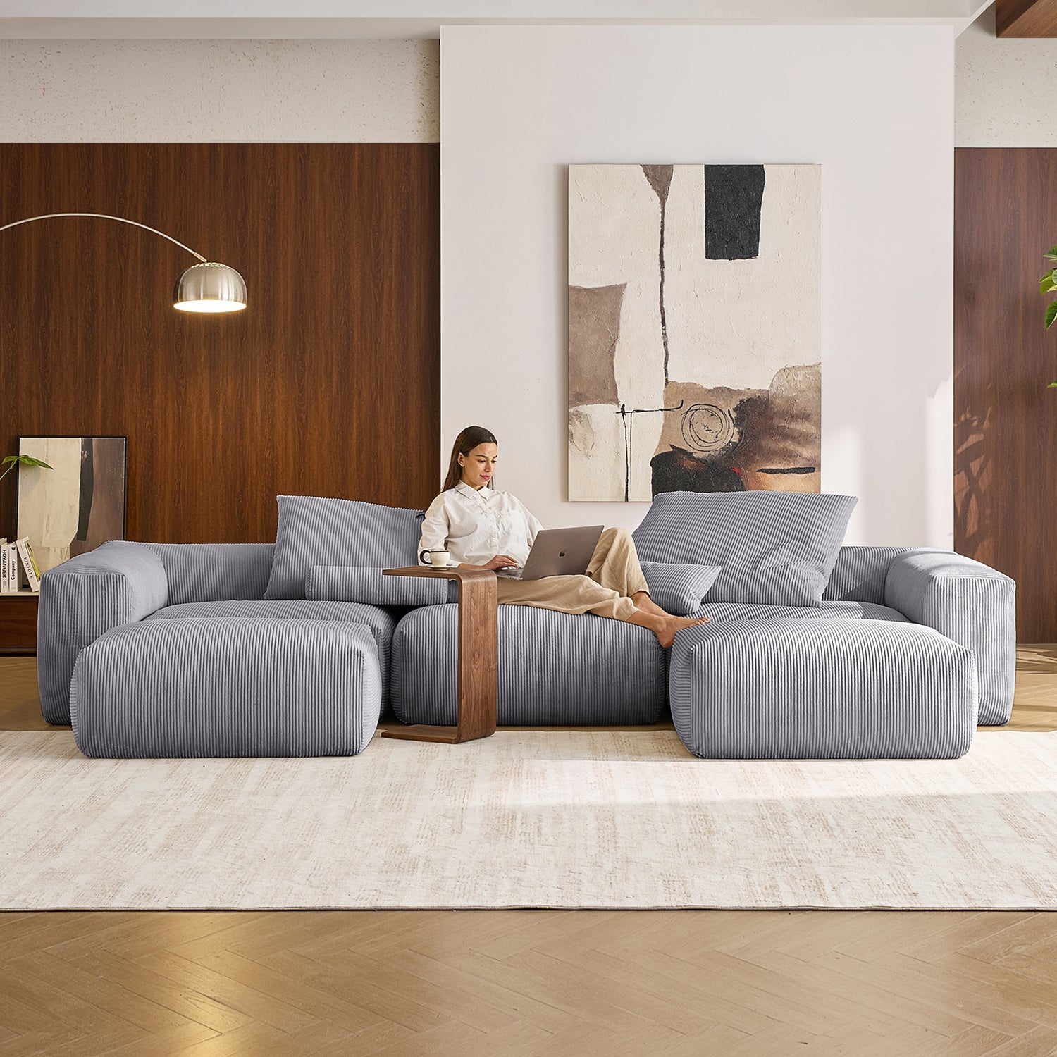 Image of a woman sitting on a cord-velour sofa sectional by Mario Capasa as she uses her laptop. Her coffee is sitting on top of the wooden side table in front of her.